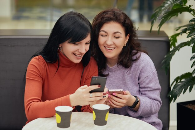 Dos hermosa mujer joven sentada en la cafetería tomando café y mirando el teléfono móvil, se ven felices, disfrutando de pasar tiempo juntos, damas con expresión positiva.