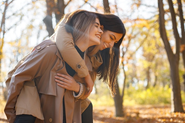 Dos hermosas amigas pasando tiempo juntas. Dos jóvenes hermanas sonrientes caminando en el parque de otoño y abrazándose. Chicas morenas y rubias con abrigos.