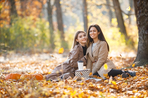 Dos hermosas amigas pasando tiempo en una manta de picnic en el césped. Dos jóvenes hermanas sonrientes haciendo picnic comiendo croissant en el parque de otoño. Chicas morenas y rubias con abrigos.