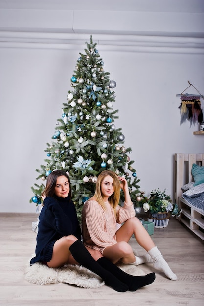 Dos hermosas amigas visten polainas de suéter cálido y calentadores de piernas contra el árbol de año nuevo con decoración navideña retrato de vacaciones de estudio de chica rubia y morena