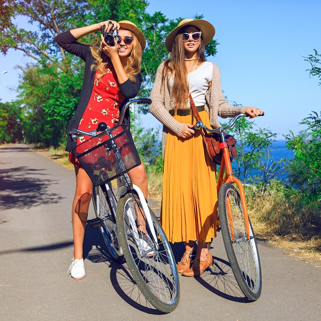 Dos hermosas chicas amigas volviéndose locas, divirtiéndose y con agradables emociones positivas, caminando con bicicletas retro, saludando. vistiendo elegantes trajes vintage, sosteniendo una cámara retro.