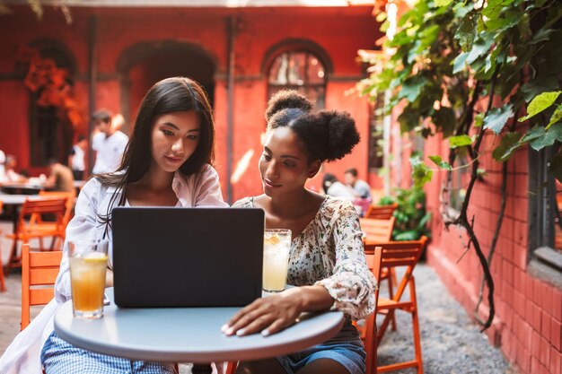 Dos hermosas chicas cuidadosamente usando una computadora portátil mientras pasan tiempo con cócteles juntas en el acogedor patio de la cafetería