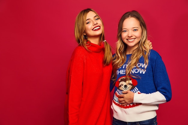 Dos hermosas chicas guapas sonrientes mirando a la cámara. Mujeres de pie en elegantes suéteres calientes de invierno sobre fondo rojo. Navidad, Navidad, concepto