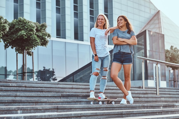 Dos hermosas chicas hipster paradas en escalones con monopatín en el fondo del rascacielos.