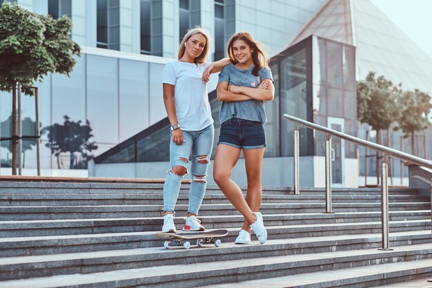Dos hermosas chicas hipster paradas en escalones con monopatín en el fondo del rascacielos.