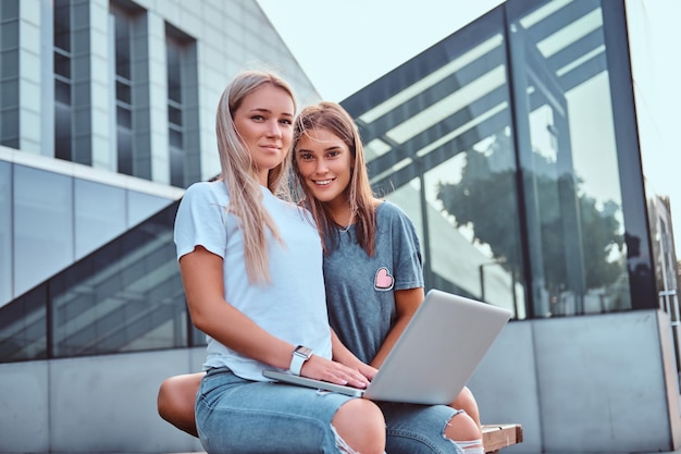 Dos hermosas chicas hipster sentadas en el banco con una laptop en el fondo del rascacielos.