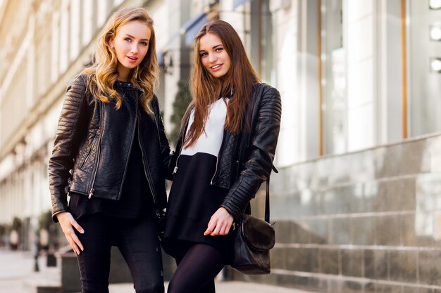 Dos hermosas chicas jóvenes posando juntos al aire libre. Estado de ánimo urbano de estilo de vida. Fondo del centro de la ciudad. Mejores amigos con traje negro casual de otoño.