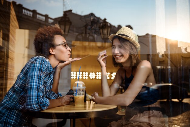 Dos hermosas chicas jóvenes sonriendo, hablando, descansando en la cafetería. Disparo desde afuera.