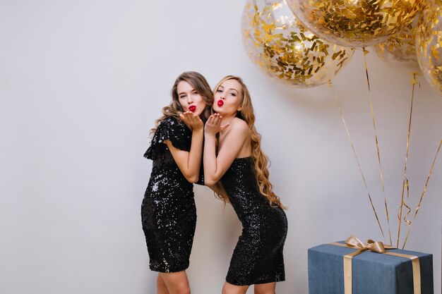 Dos hermosas chicas en vestidos negros similares posando con expresión de cara de besos en la fiesta de cumpleaños. Señora europea de pelo largo de pie junto a globos y regalos, enviando beso aéreo.