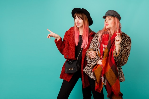 Dos hermosas mujeres con elegantes abrigos de piel sintética y bufanda de lana posando en la pared turquesa