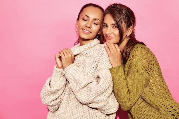 Dos hermosas mujeres hermosas sonrientes sexy. Mujeres calientes de pie y abrazándose en elegantes suéteres blancos y verdes, en la pared de color rosa.
