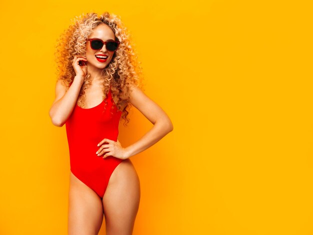 Dos hermosas mujeres hipster sonrientes sexy en trajes de baño de verano rojo Modelos de moda con peinado de rizos afro divirtiéndose en el estudio Mujer caliente aislada en amarillo En gafas de sol