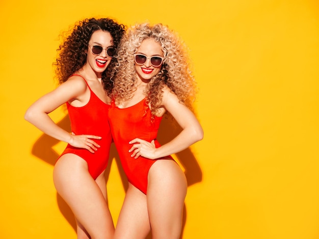 Dos hermosas mujeres hipster sonrientes sexy en trajes de baño de verano rojo modelos de moda con peinado de rizos afro divirtiéndose en el estudio mujer caliente posando junto a la pared amarilla en gafas de sol