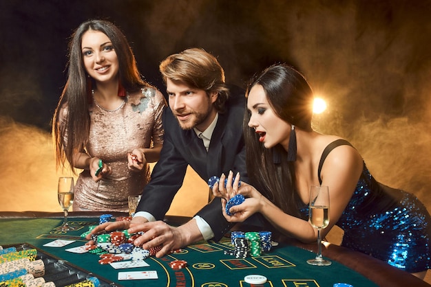 Dos hermosas mujeres y un joven juegan en la mesa de póquer en el casino, se centran en el hombre y la morena. Pasión, cartas, fichas, alcohol, dados, juegos de azar, casino: es entretenimiento. Juego de cartas divertido y peligroso para mo