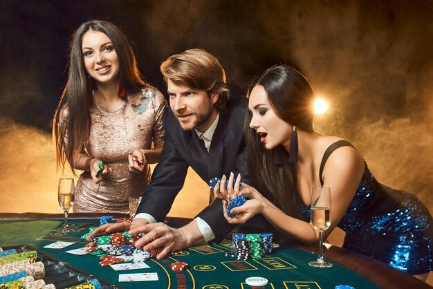 Dos hermosas mujeres y un joven juegan en la mesa de póquer en el casino, se centran en el hombre y la morena. Pasión, cartas, fichas, alcohol, dados, juegos de azar, casino: es entretenimiento. Juego de cartas divertido y peligroso para mo