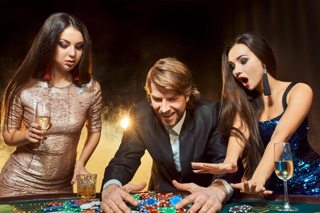 Dos hermosas mujeres y un joven juegan en la mesa de póquer en el casino, se centran en el hombre y la morena. Pasión, cartas, fichas, alcohol, dados, juegos de azar, casino: es entretenimiento. Juego de cartas divertido y peligroso para mo