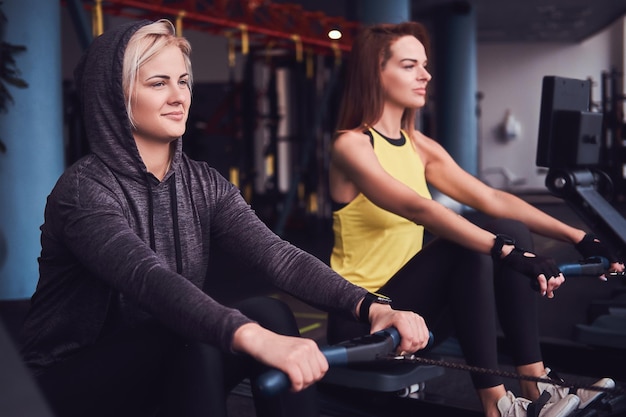 Dos hermosas mujeres jóvenes practicando remo en el moderno gimnasio