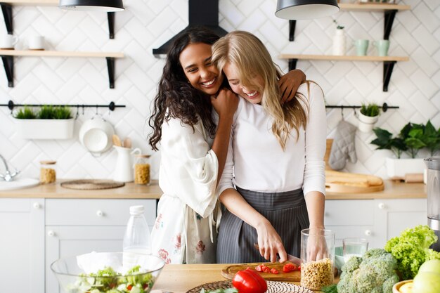 Dos hermosas mujeres jóvenes preparan un desayuno saludable y se abrazan cerca de la mesa llena de verduras frescas en la moderna cocina blanca