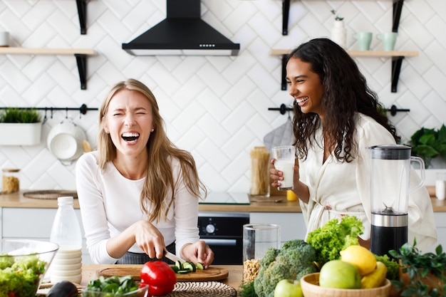 Dos hermosas mujeres jóvenes preparan un desayuno saludable y se ríen sinceramente cerca de la mesa llena de verduras frescas en la moderna cocina blanca.