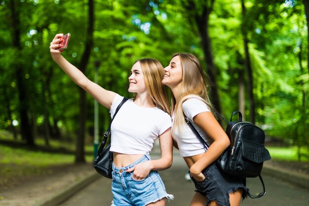 Dos hermosas mujeres jóvenes toman selfie en el teléfono en el parque soleado. Novias.