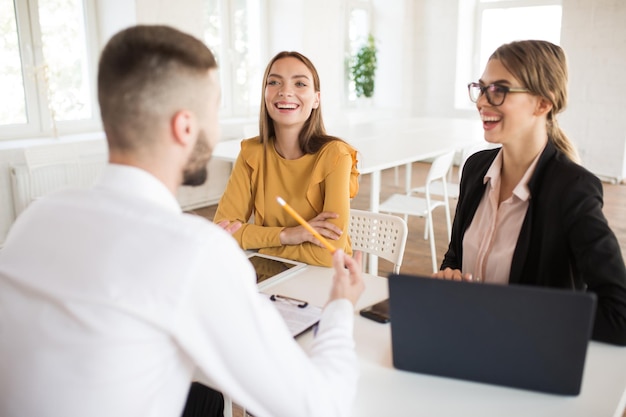 Foto gratuita dos hermosas mujeres de negocios hablando felizmente con un candidato masculino para el trabajo jóvenes empleadores sonrientes que pasan una entrevista de trabajo en una oficina moderna