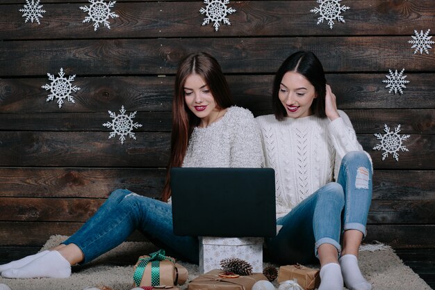 Dos hermosas mujeres sentadas en el suelo con un ordenador portátil, entre los regalos de Navidad