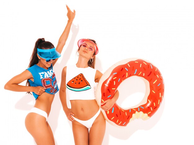 Dos hermosas mujeres sexy sonrientes en calzoncillos de verano blanco y tema. Chicas con gafas de sol, visera transparente. Modelos positivos divirtiéndose con el colchón inflable donut lilo. Aislado