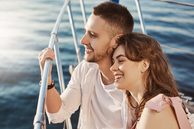 Dos hermosas personas casadas enamoradas, sonriendo ampliamente mientras están sentados en la proa del barco y sosteniendo la barandilla. Una pareja de adultos jóvenes en una relación comparte historias sobre sus ex.