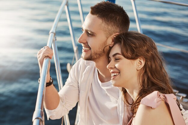 Dos hermosas personas casadas enamoradas, sonriendo ampliamente mientras están sentados en la proa del barco y sosteniendo la barandilla. Una pareja de adultos jóvenes en una relación comparte historias sobre sus ex.