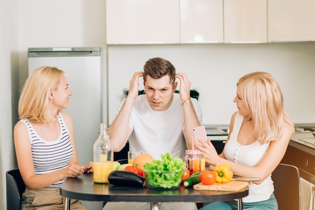 Dos hermosas rubias caucásicas escuchando a su amigo cuenta una historia interesante. tres amigos sentados a la mesa en la cocina, bebiendo batidos de frutas y verduras. personas, concepto de comida sana.