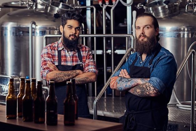 Dos hipsters interraciales hombres barbudos con camisas y delantales trabajando en la cervecería.