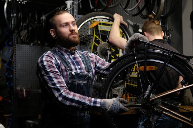 Dos hombres guapos y elegantes que trabajan con una bicicleta en un taller de reparación. Los trabajadores reparan y montan bicicletas en un taller.