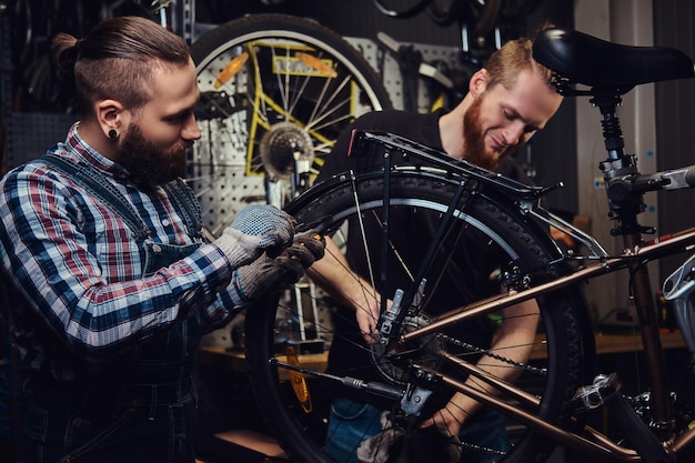 Dos hombres guapos y elegantes que trabajan con una bicicleta en un taller de reparación. Los trabajadores reparan y montan bicicletas en un taller.