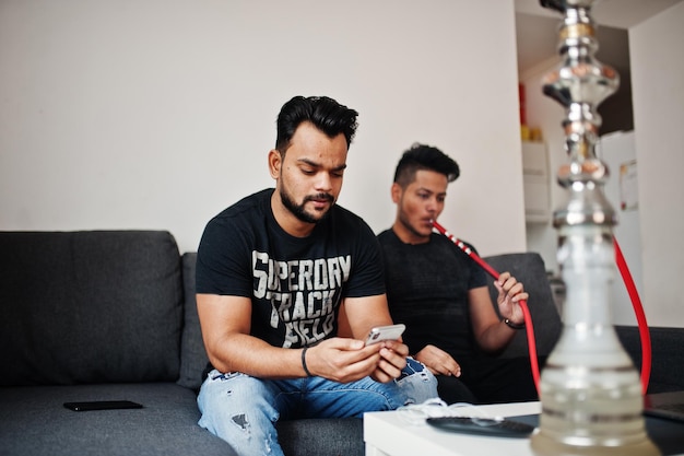 Foto gratuita dos hombres indios guapos y de moda relajándose en la habitación y fumando narguile uno de ellos mirando el teléfono móvil