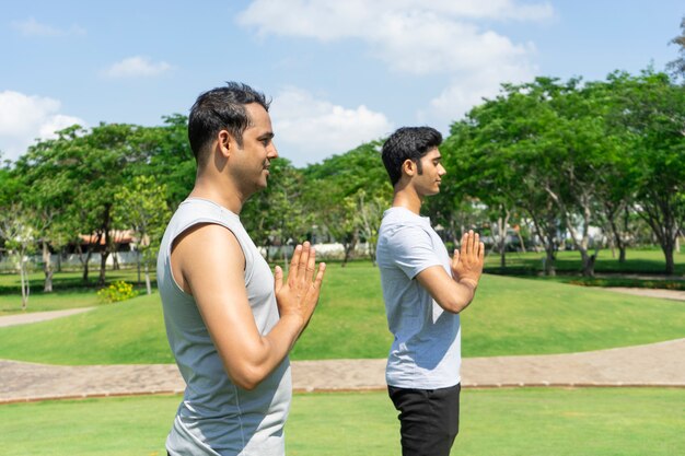 Dos hombres indios serenos meditando y manteniendo las manos juntas en el parque con árboles verdes