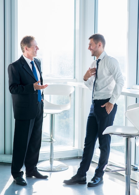 Dos hombres de negocios sonrientes de pie junto a la ventana con conversación en la oficina