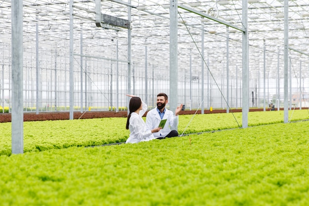 Dos investigaciones hombre y mujer examinan la vegetación con una tableta en un invernadero blanco
