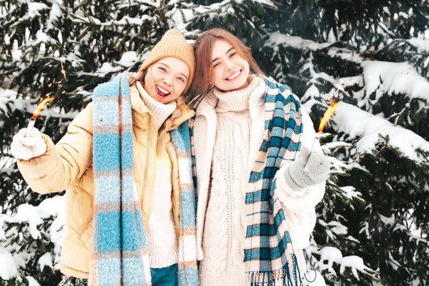Dos joven hermosa mujer hipster sonriente en bufandas y ropa de abrigo de moda. Mujeres despreocupadas posando en la calle en el parque. Modelos puros positivos abrazándose. Sosteniendo un palo brillante ardiente