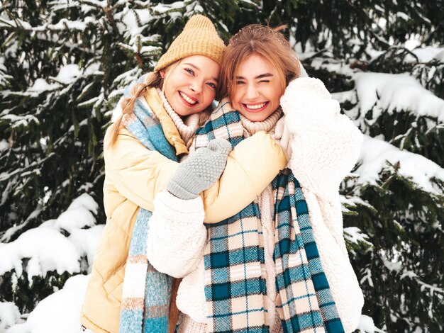 Dos joven hermosa mujer hipster sonriente en ropa de abrigo de moda y bufandas. Mujeres despreocupadas posando en la calle en el parque. Modelos puros positivos divirtiéndose en la nieve. Disfrutando de los momentos invernales. Concepto de navidad