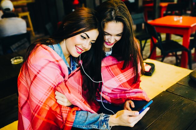 Dos joven y hermosa niña sentada en la mesa escuchando música con un teléfono inteligente