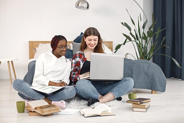 Dos jóvenes adolescentes sentadas en un piso cerca de la cama estudiando y usando una computadora portátil