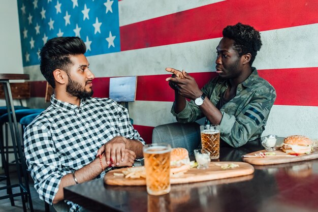 Dos jóvenes alegres bebiendo cerveza y comen hamburguesas en el bar americano moderno.