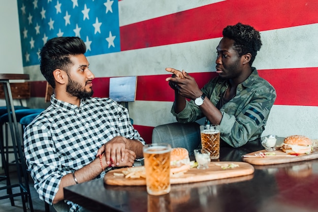 Dos jóvenes alegres bebiendo cerveza y comen hamburguesas en el bar americano moderno.