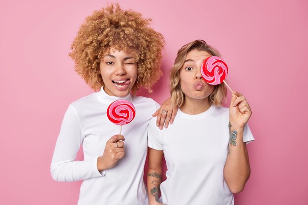Dos jóvenes amigas se divierten sosteniendo caramelos en palos haciendo muecas a la cámara vestidas con ropa blanca casual aislada sobre fondo rosa Mujeres amistosas con deliciosas piruletas