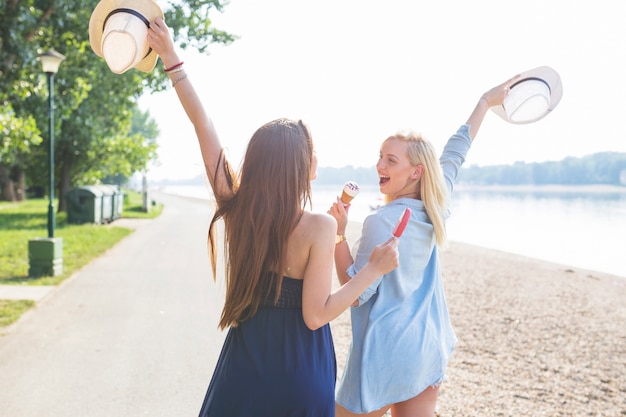 Dos jóvenes amigas sosteniendo sombrero disfrutando de helado en la playa