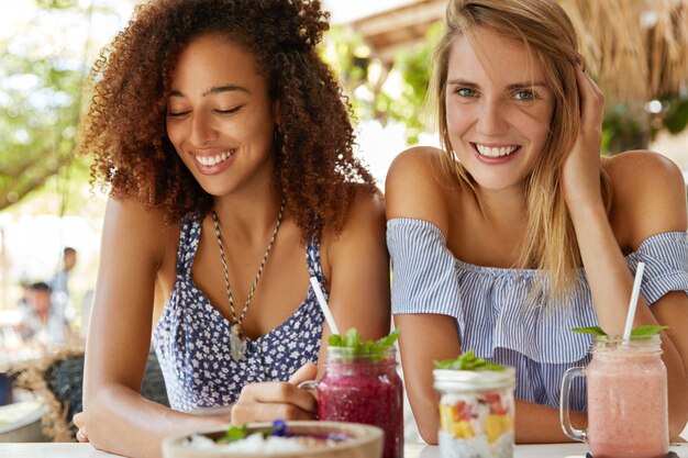 Dos jóvenes amigas tienen sonrisas positivas vestidas con ropa de verano, comen postres exóticos y beben batidos, disfrutan del tiempo de recreación en el interior de la cafetería de la terraza, estando de buen humor.