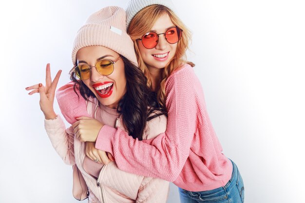 Dos jóvenes amigos alegres en perfecto estado de ánimo teniendo tiempo juntos sobre fondo blanco aislado. Chicas positivas con suéteres y pulóver rosa acogedores, sombrero de moda, gafas lindas.