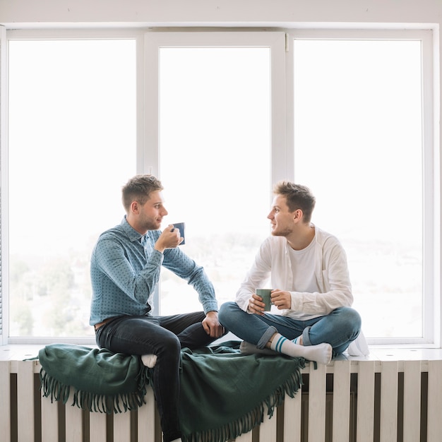 Dos jóvenes amigos discutiendo entre sí sosteniendo la taza de café sentado cerca de la ventana