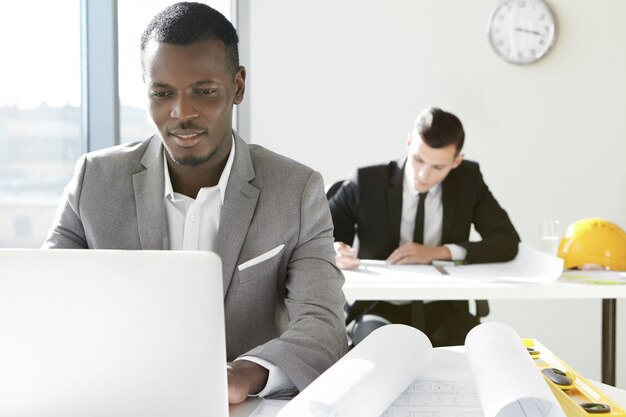 Dos jóvenes arquitectos de la empresa de ingeniería que trabajan en la oficina. Diseñador africano desarrollando un nuevo proyecto de construcción usando una computadora portátil, sentado en un escritorio con rollos y regla.