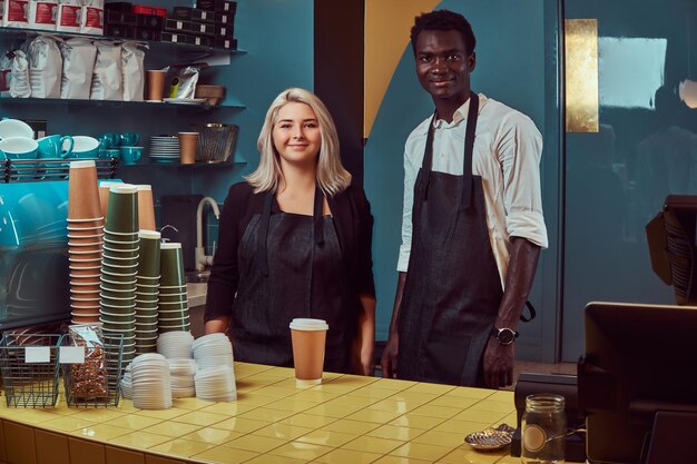 Dos jóvenes baristas multirraciales en delantales de pie en su cafetería de moda.